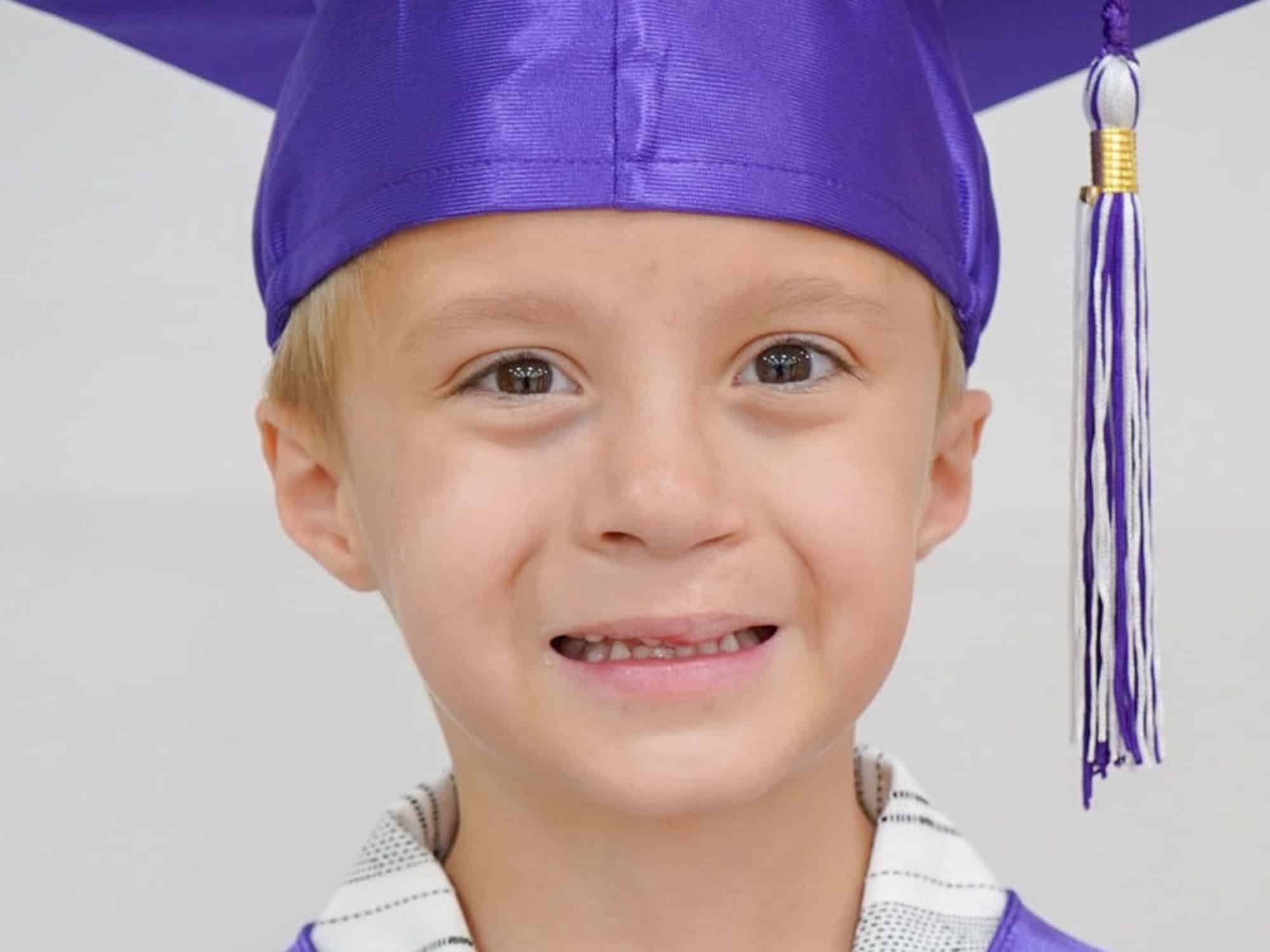 Smiling child wearing a purple graduation cap and gown with a tassel against a plain background, showcasing a joyful and proud expression.