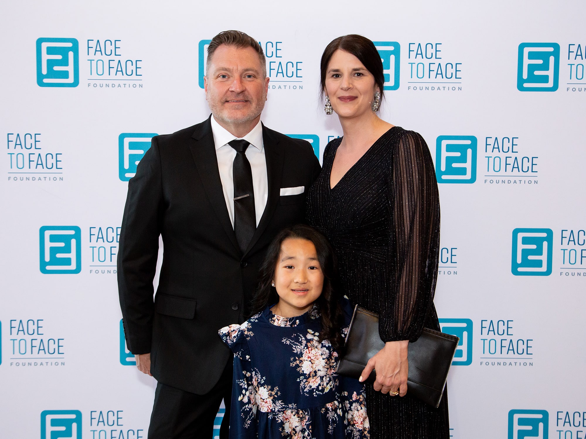 Three people dressed formally pose in front of a "Face to Face Foundation" backdrop at an event.