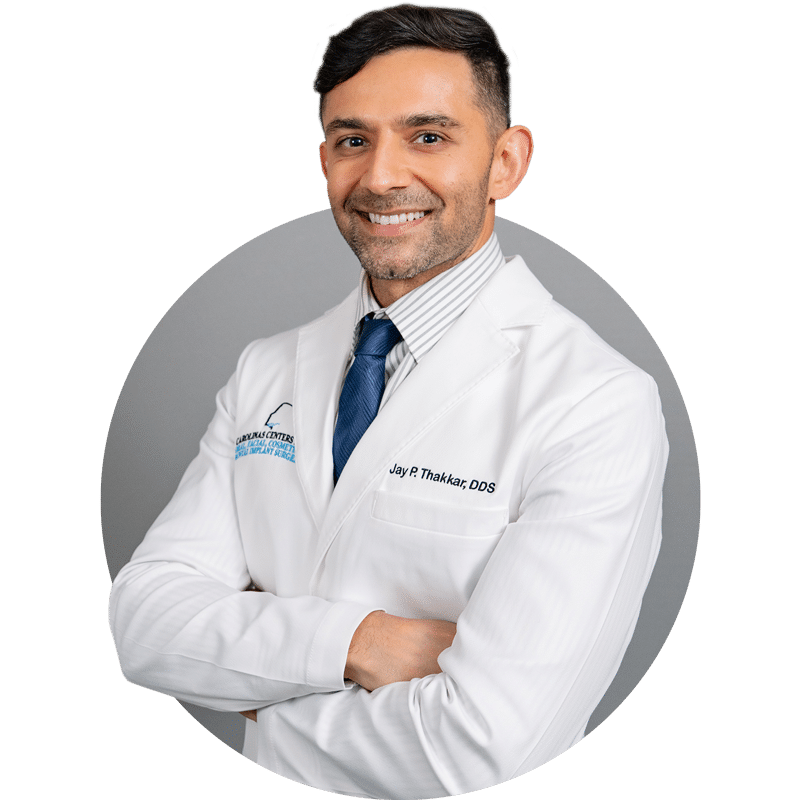 A person in a white coat with a blue tie smiles, standing against a gray background, signifying a professional healthcare setting.