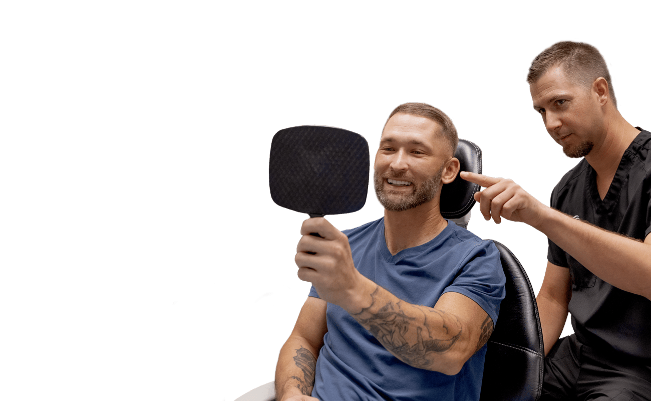 A person sits in a dental chair smiling at a hand mirror, while another person points, likely discussing dental results.