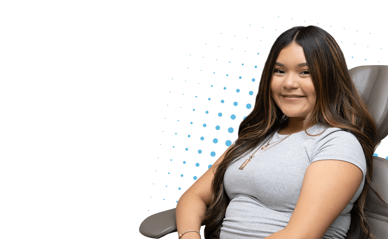 A person with long hair wearing a gray shirt smiles, sitting indoors against a dotted blue pattern background.