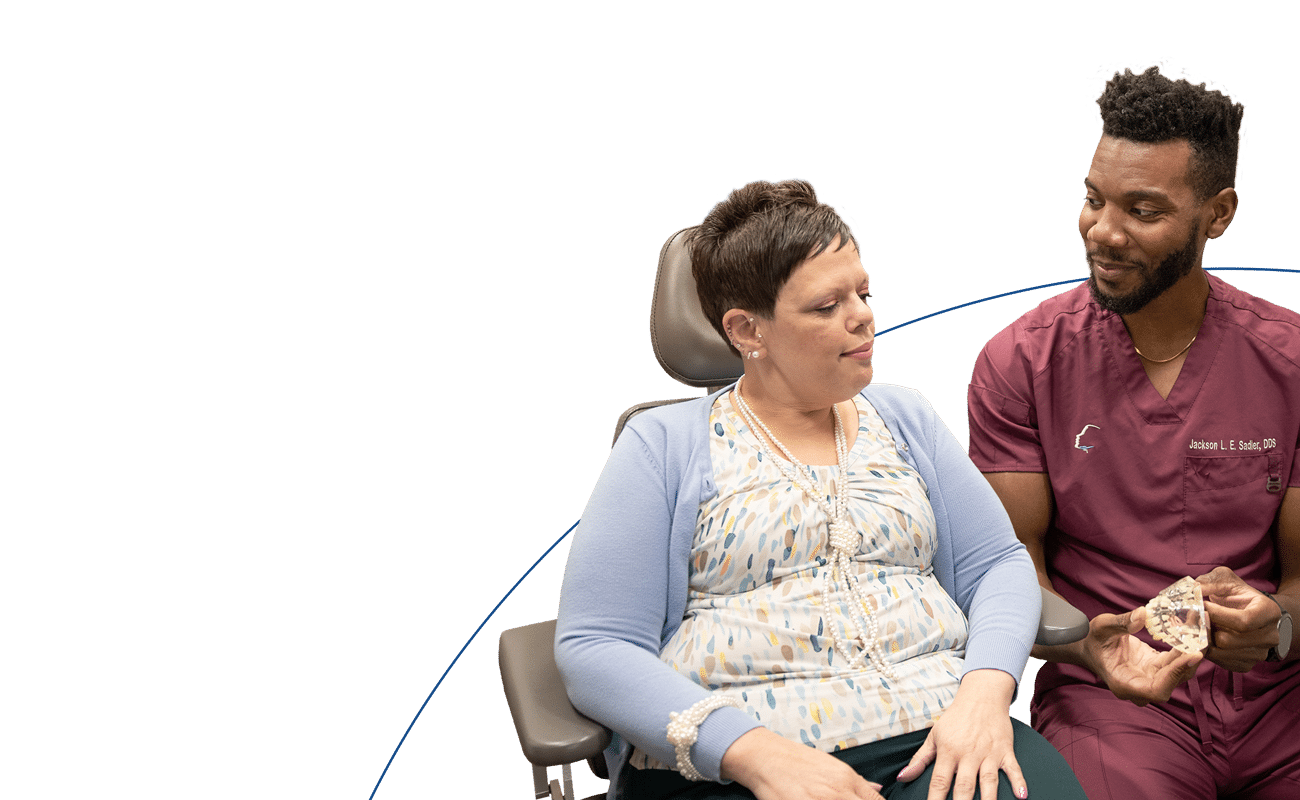 A person in scrubs shows a dental model to a seated person against a blue and white background, suggesting a dental consultation.