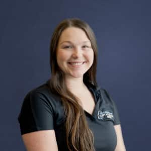 A person with long hair smiles in front of a plain, dark blue background, wearing a black shirt with text partially visible.