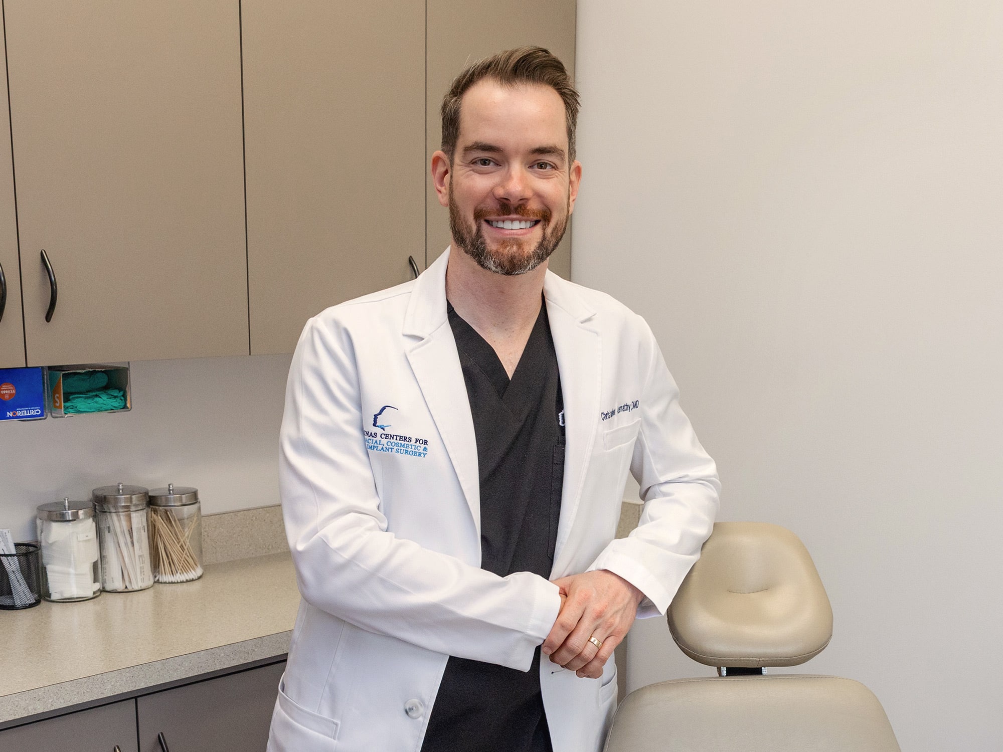 A person in a medical office wearing a white coat smiles while standing beside an exam chair, surrounded by clinic supplies.
