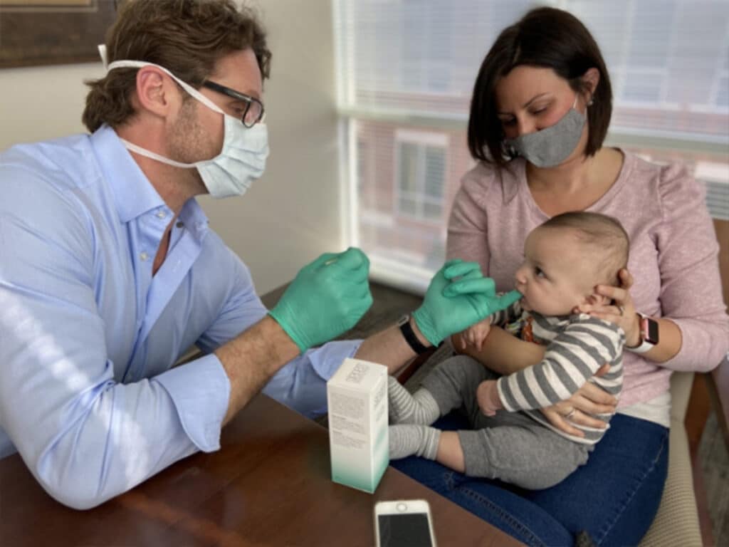 A person wearing gloves and a mask checks a baby's mouth while another person holds the baby, both wearing masks indoors.