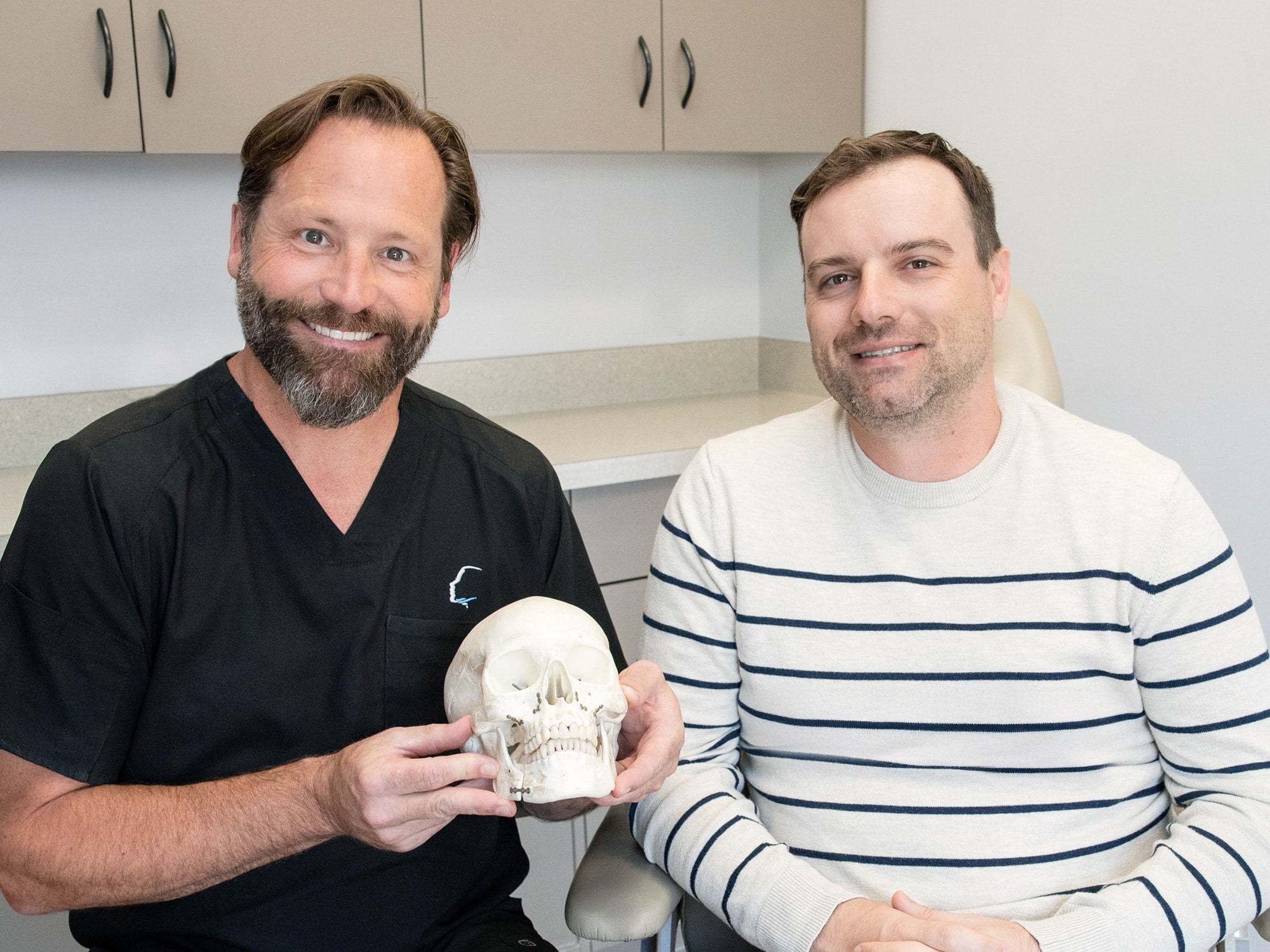 Two people smiling, seated indoors, one holding a skull model. Background includes cabinets and a countertop. Casual, friendly atmosphere.