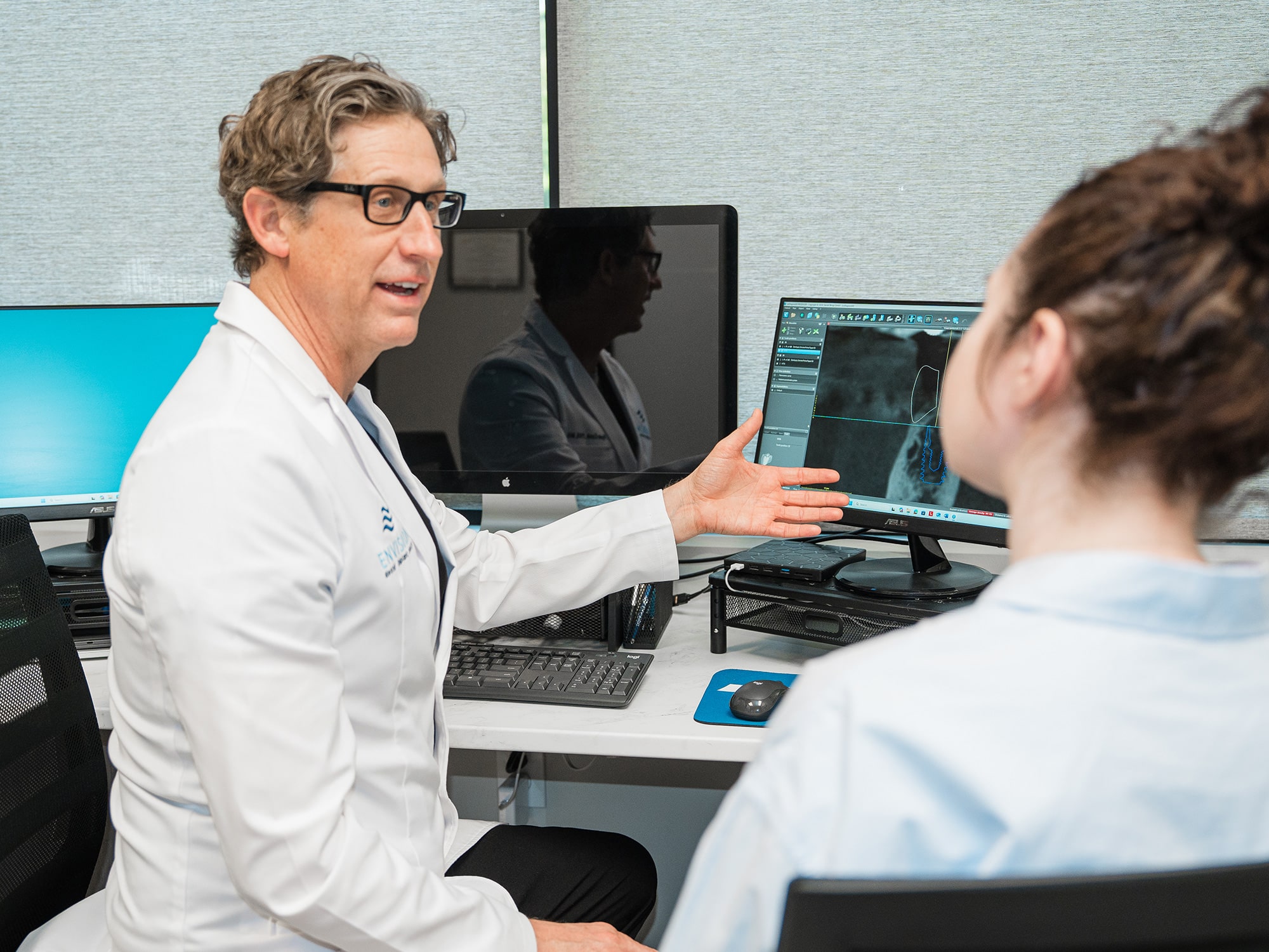 A person in a lab coat discusses medical imaging on computer screens with another person in an office setting.