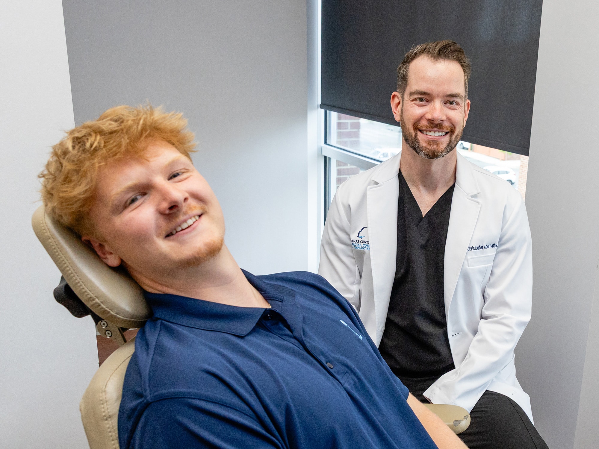 A person in medical attire sits beside a smiling person reclining in a chair, in a clinical setting with window light.