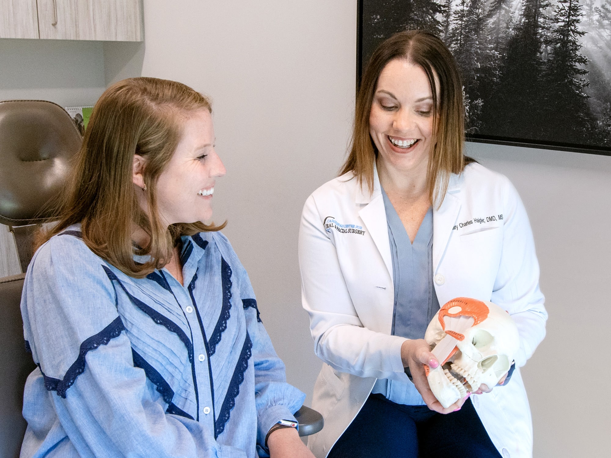 Two people in a medical office, one wearing a white coat, discuss a human skull model, smiling while seated near a dental chair.