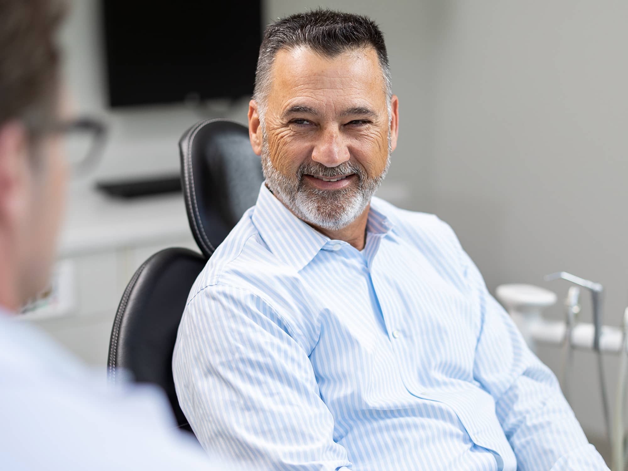A person with a beard smiles while sitting in a chair, engaged in conversation with another person. The setting appears clinical.