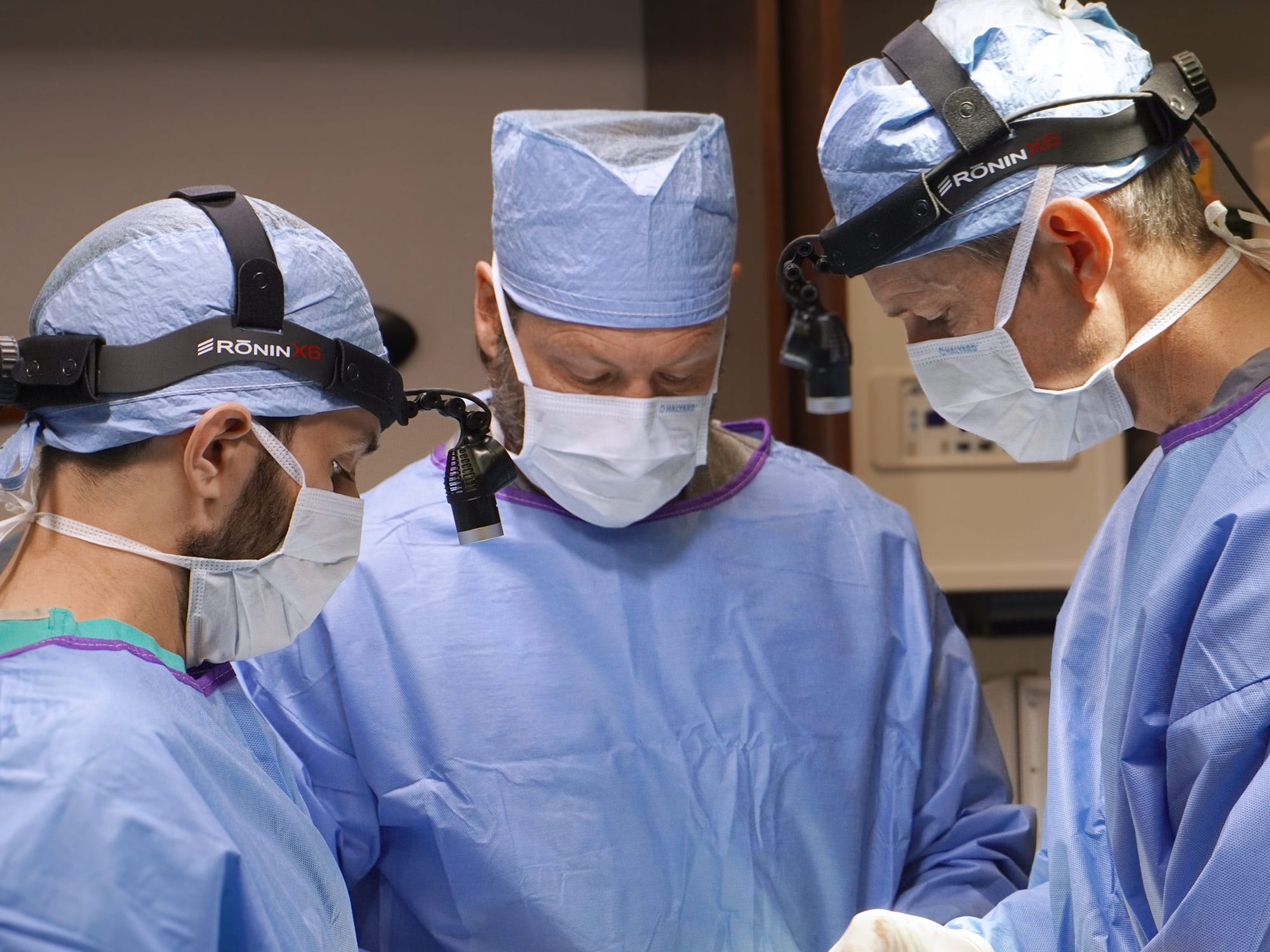 Three people in surgical attire focus on a medical procedure. They wear masks, blue gowns, and headlamps, concentrating intently on their work.
