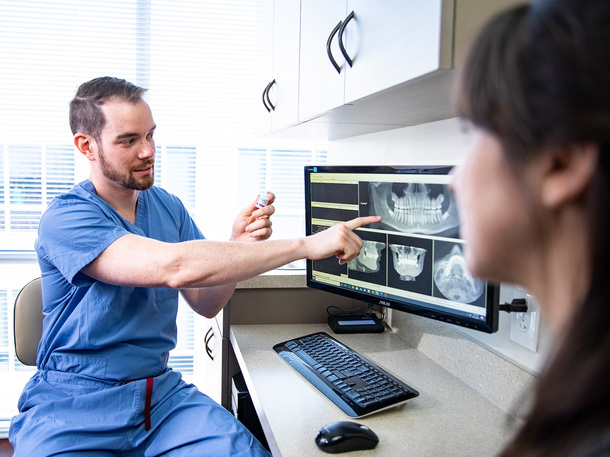 A person in scrubs discusses dental X-rays with another person in a dental office, pointing at the monitor, illustrating patient consultation.