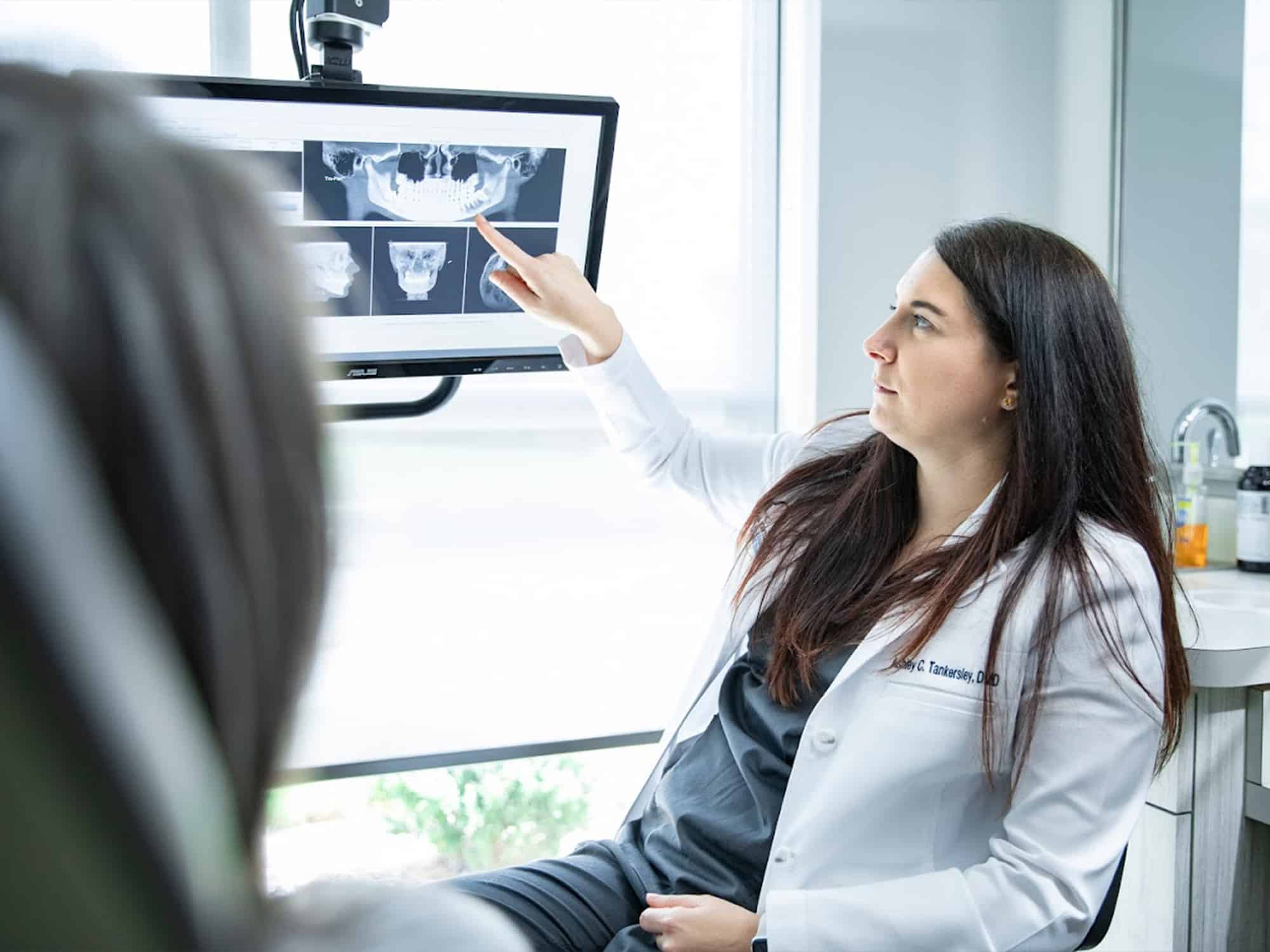 A person in a white coat points at dental X-rays on a computer screen in a bright, modern, medical office.