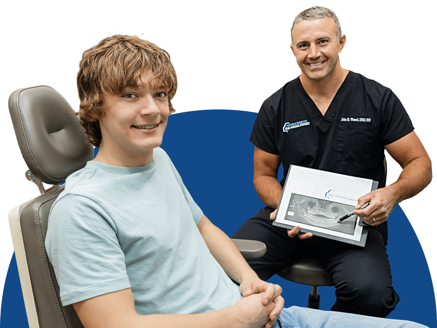 A person in scrubs holds a dental X-ray while seated next to a smiling patient in a dental chair.
