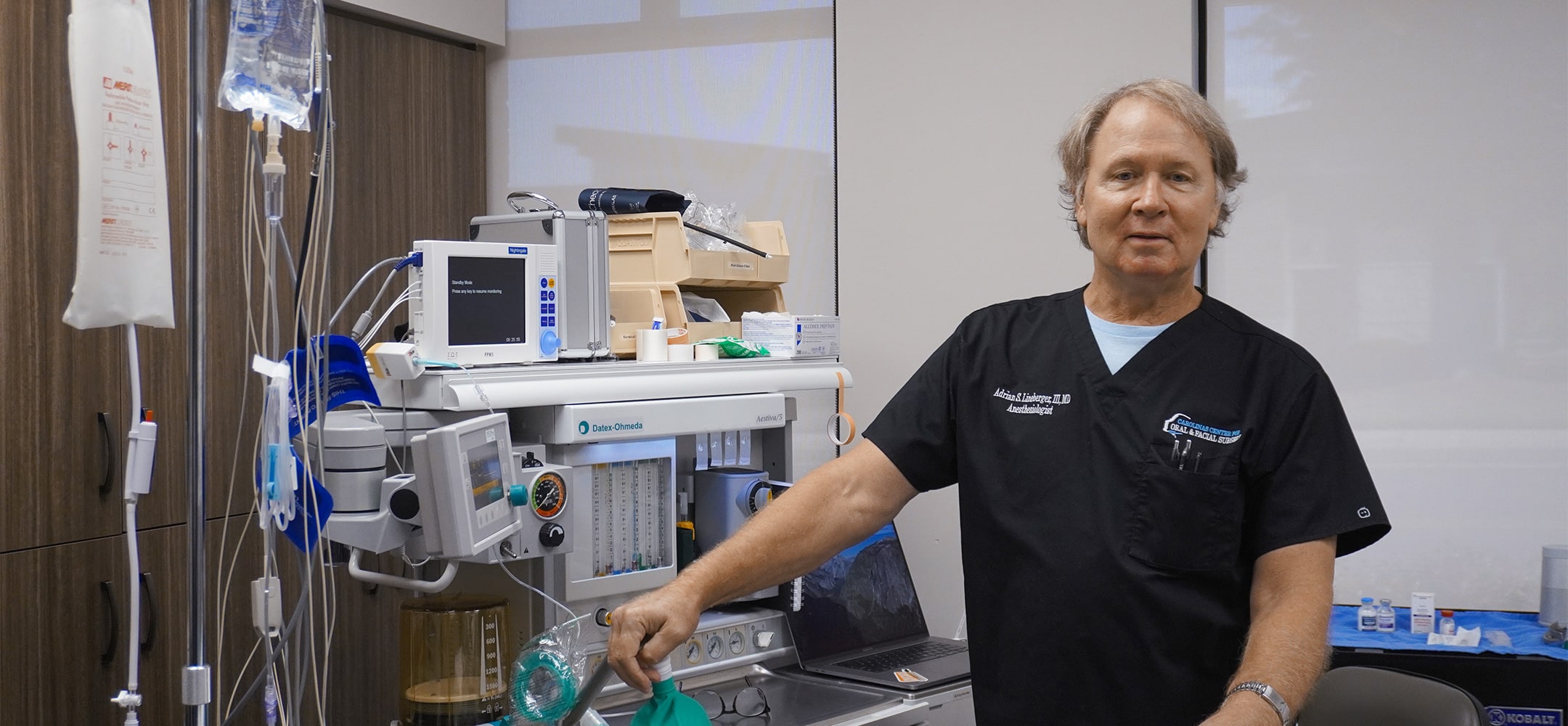 A person in medical scrubs stands beside anesthesia equipment in a clinical setting, holding a face mask.