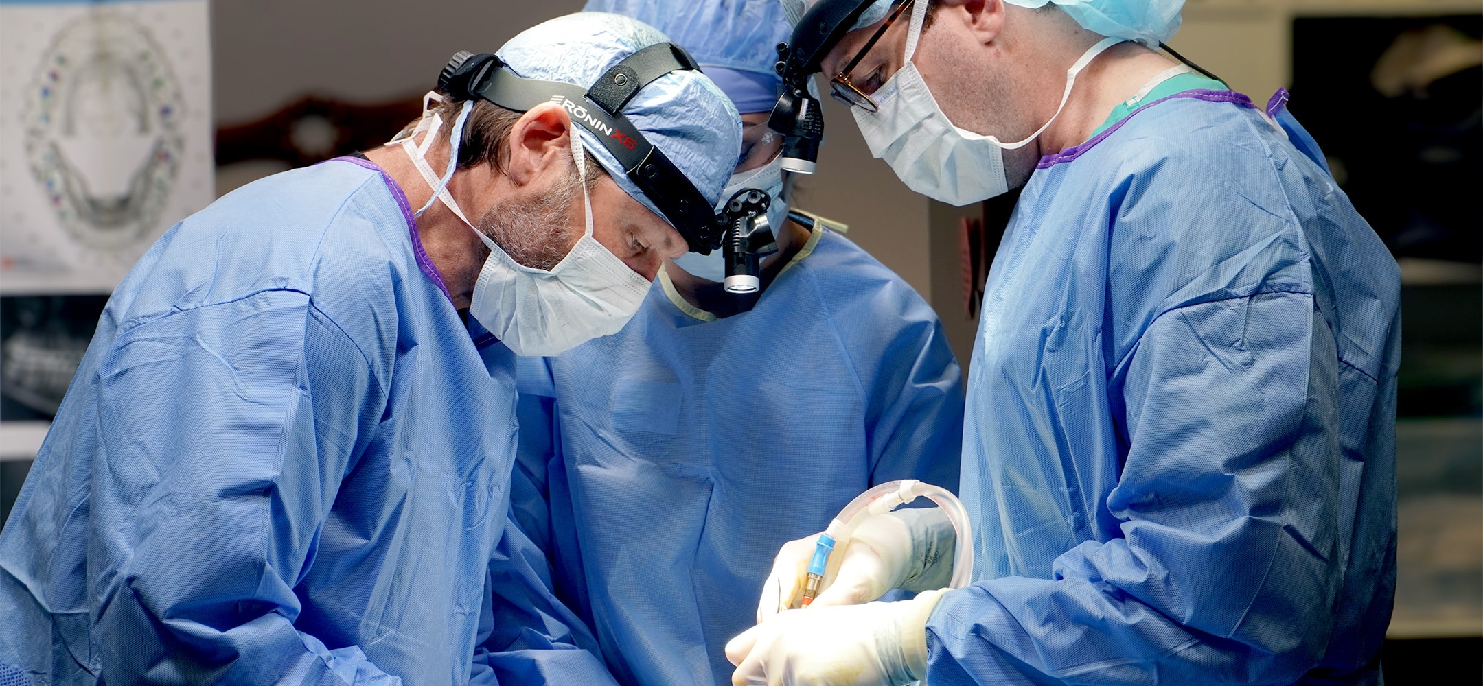 Three medical professionals in blue surgical attire focus intently on a procedure, using advanced equipment and displaying teamwork in a sterile environment.