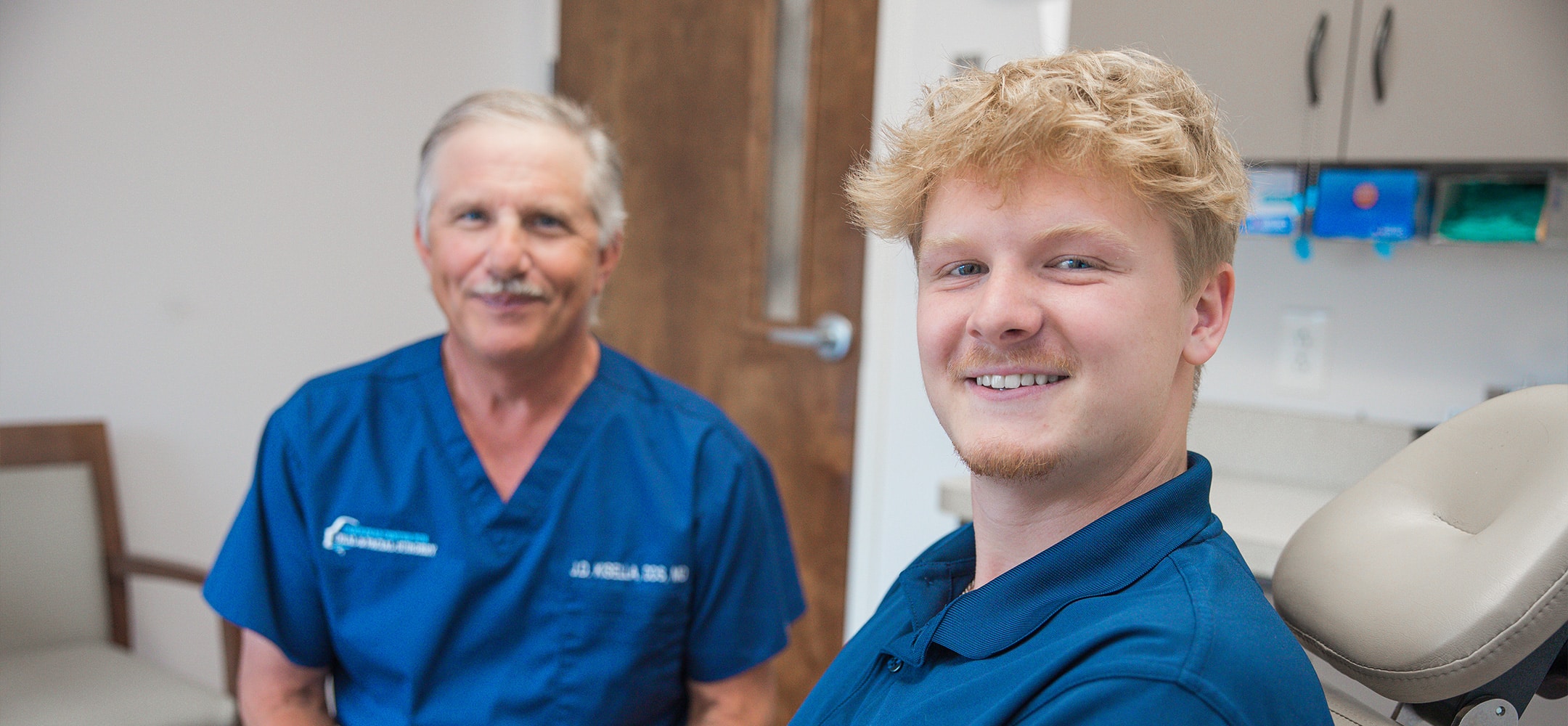Two people in blue outfits smiling in a medical office setting, seated near a chair, with cabinets in the background.
