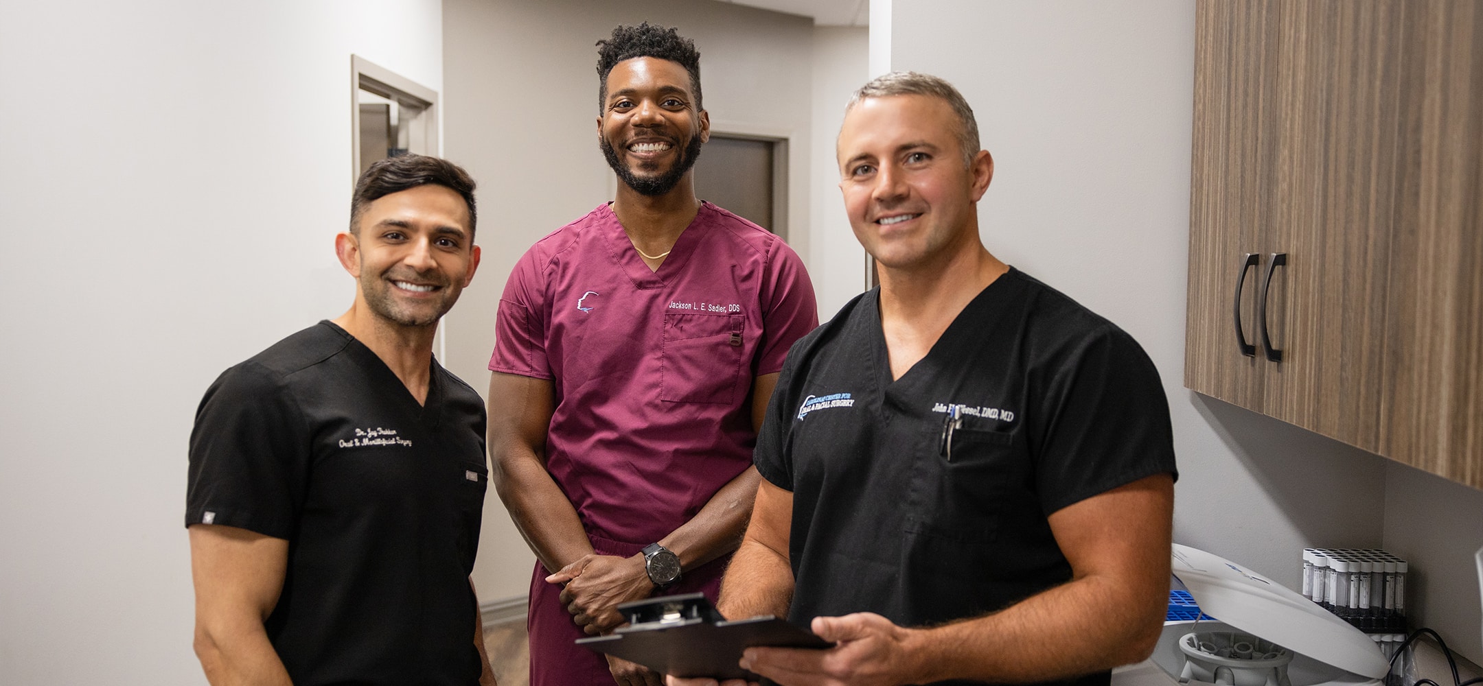 Three people in medical scrubs stand in a brightly lit room, smiling at the camera. A clipboard is held by one person.