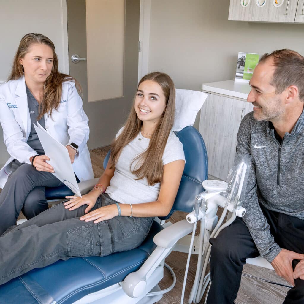 A person in a dental office converses with two others; one wears a white coat, and the patient smiles in a dental chair.