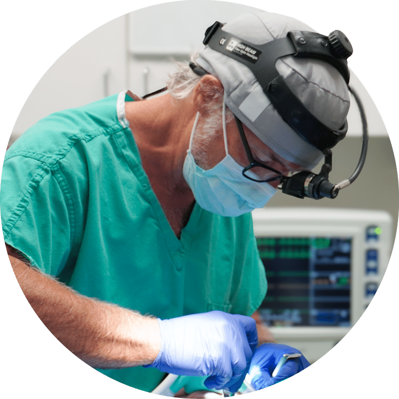 A person in surgical attire works intently during a medical procedure, with monitoring equipment visible in the background of the operating room.