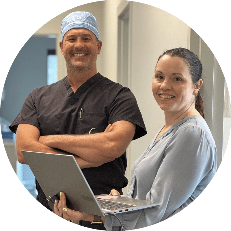 Two smiling people in a hallway, one in scrubs with a cap, the other holding a laptop, standing side by side indoors.