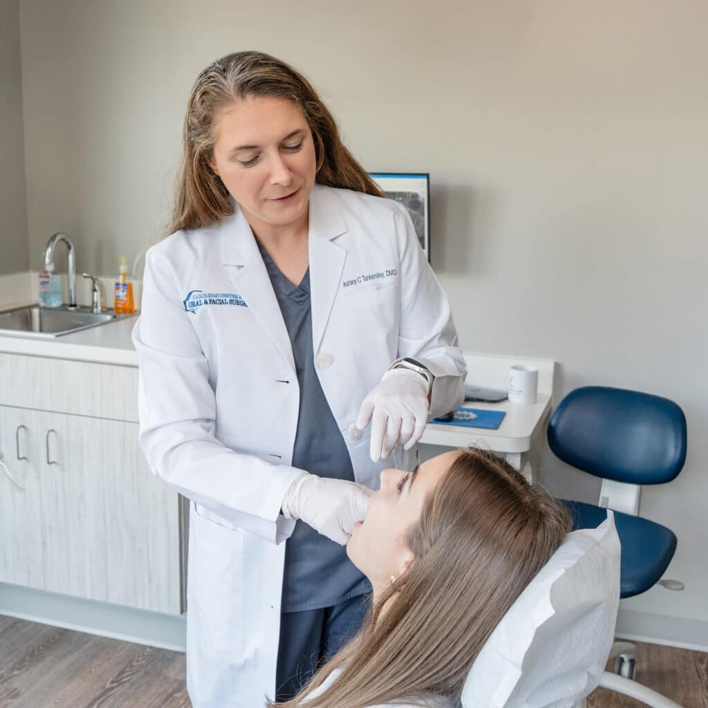 A person in a dental clinic examines another person's mouth, wearing a white coat and gloves, with dental equipment in the background.