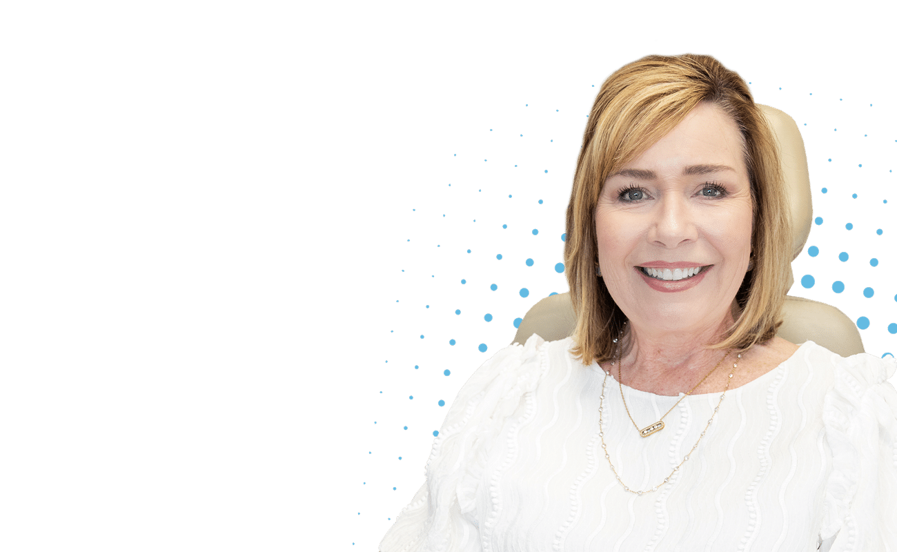 A person with light hair smiles against a dark background with blue dots, wearing a white top and a necklace.