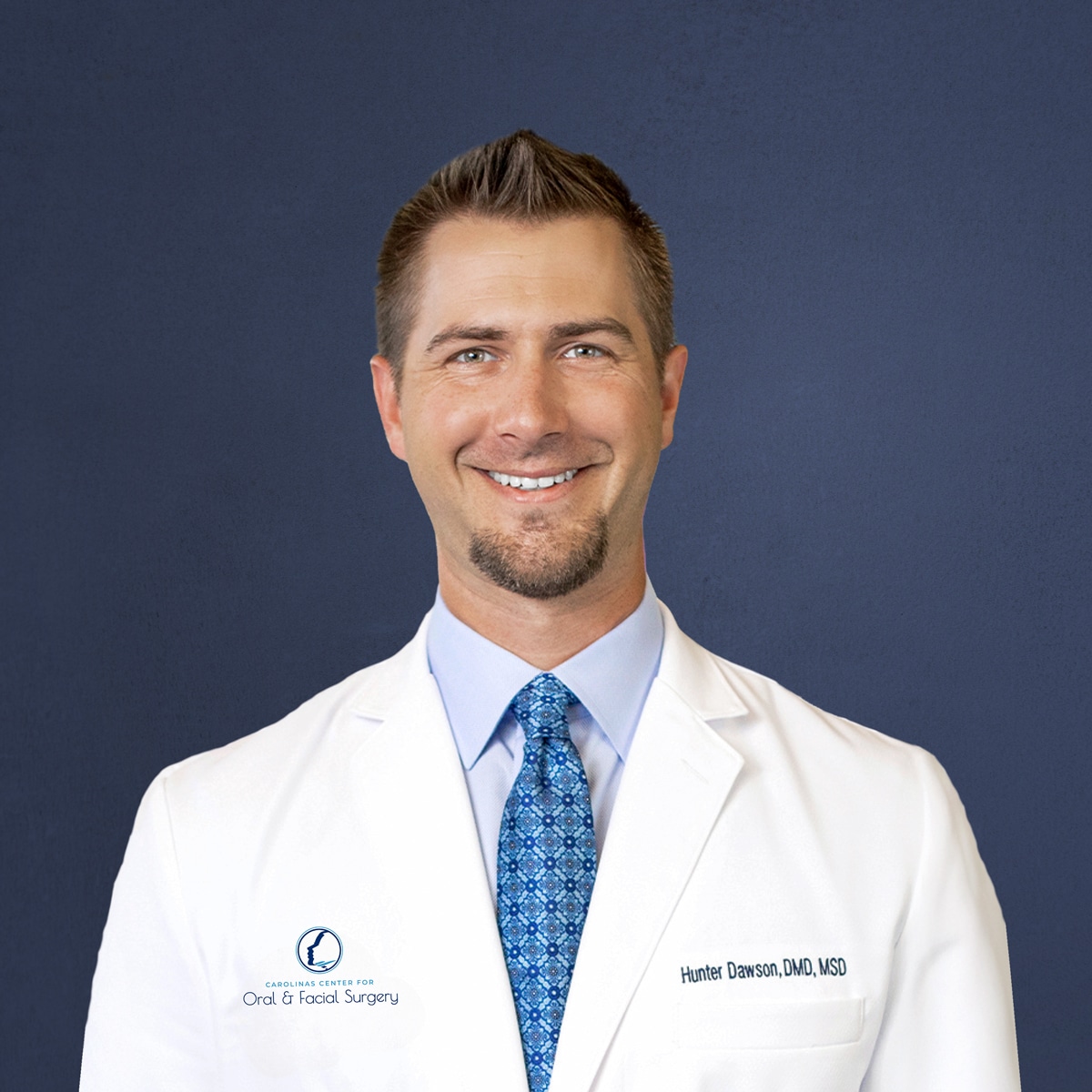 A smiling person in a white lab coat with a patterned tie against a plain dark blue background.