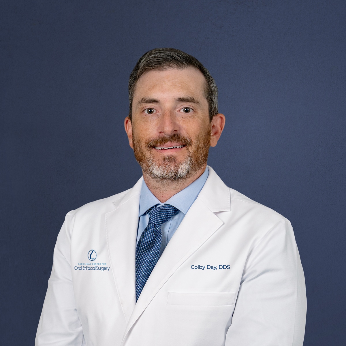A person in a white coat with "Colby Day, DDS" written, stands against a plain background, smiling slightly and wearing a blue tie.