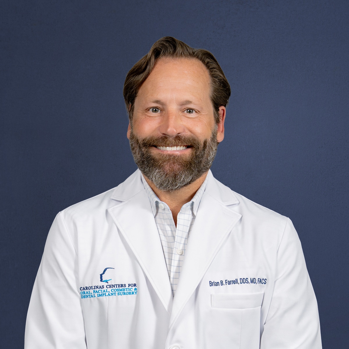 A person with a beard smiles, wearing a white coat with "CAROLINAS CENTERS" logo, against a plain blue background.