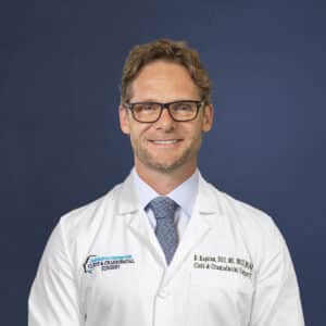 A person wearing glasses and a white lab coat with embroidered text poses smiling against a plain blue background.