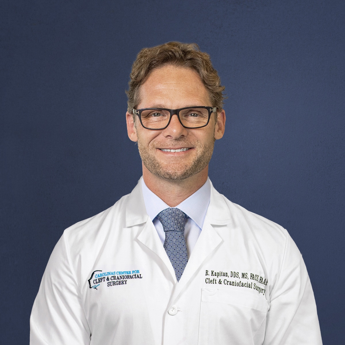 A person wearing glasses and a white lab coat with embroidered text poses smiling against a plain blue background.