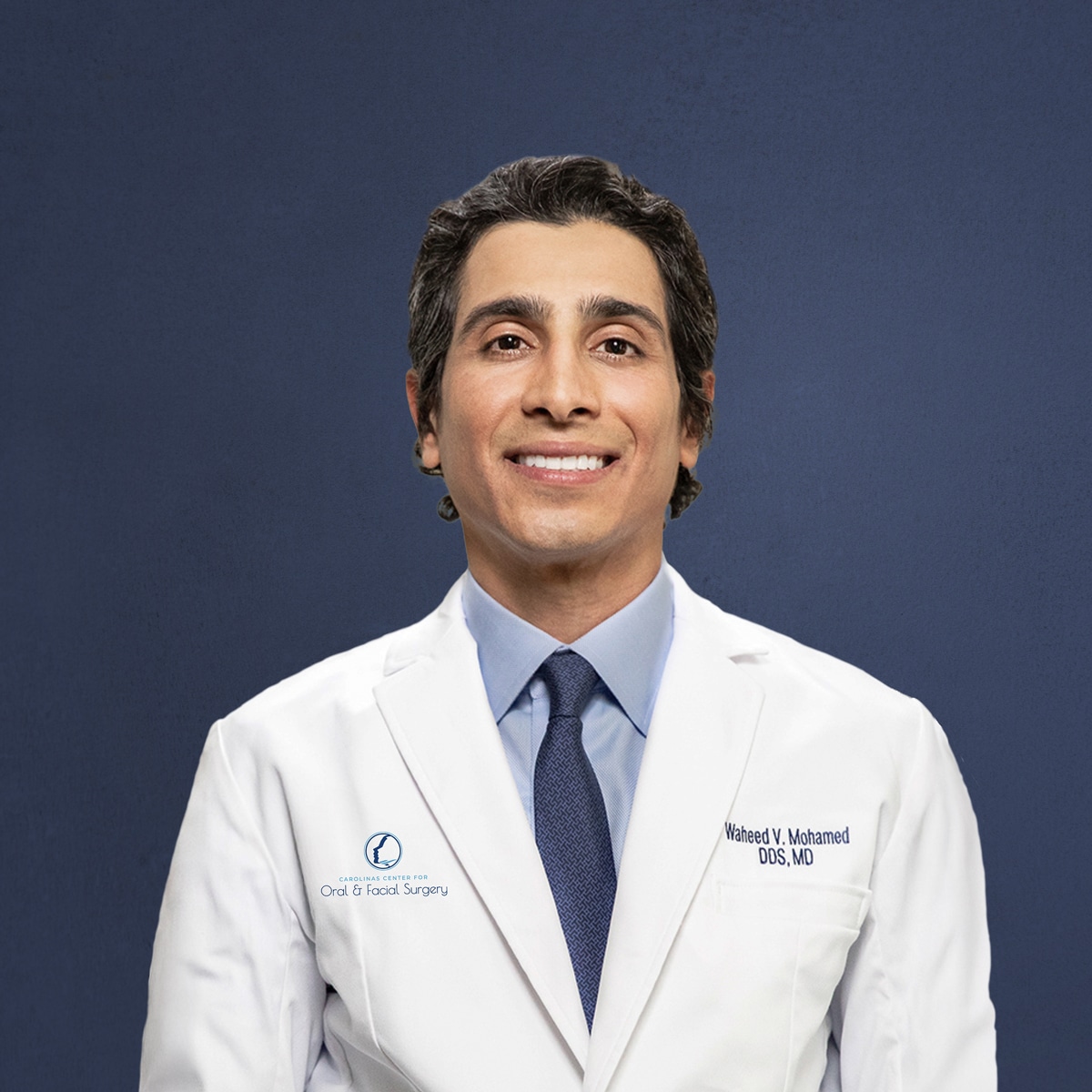 A person in a white lab coat smiles against a plain background. The coat displays medical credentials and a logo for oral and facial surgery.
