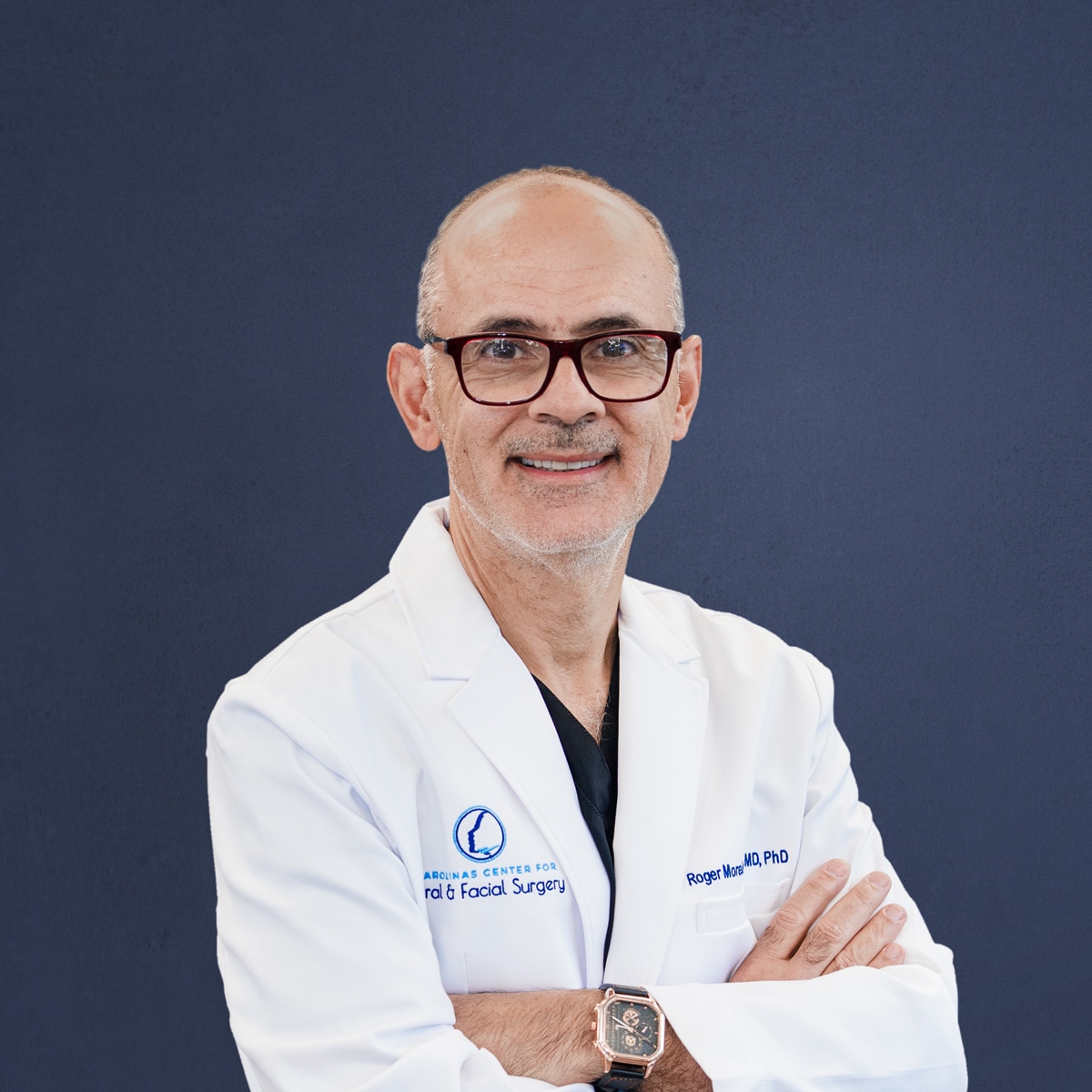 A person in a white coat with glasses and a watch stands against a blue background, smiling with crossed arms.