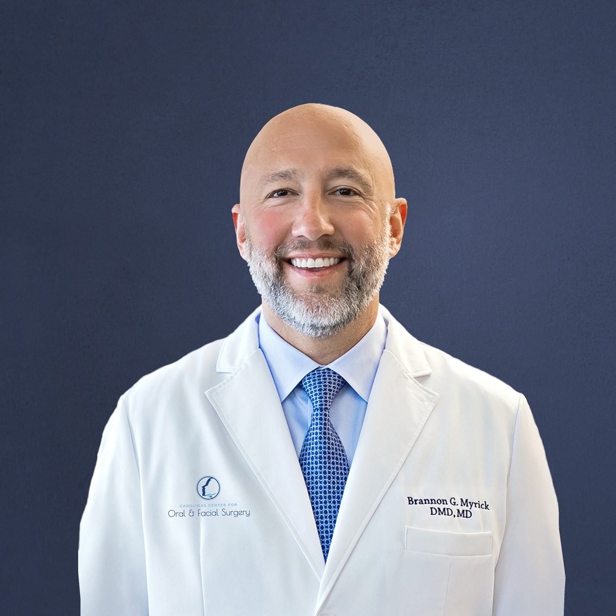 A smiling person in a white coat with a logo, against a plain blue background, wearing a blue tie.