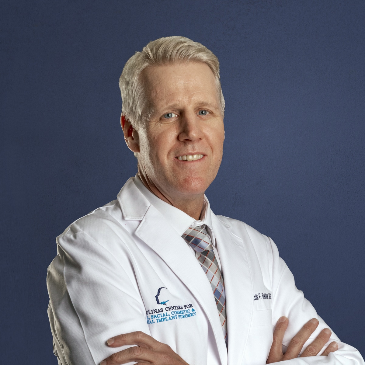 A person in a white lab coat with text, smiling, stands against a plain blue background.