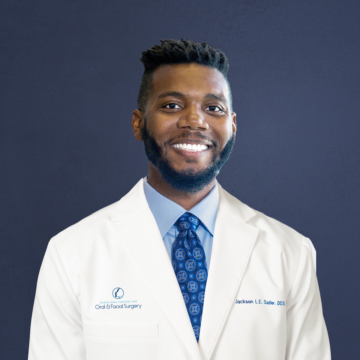 A person smiles, wearing a white coat and blue patterned tie, standing against a plain background. The coat has an embroidered logo.