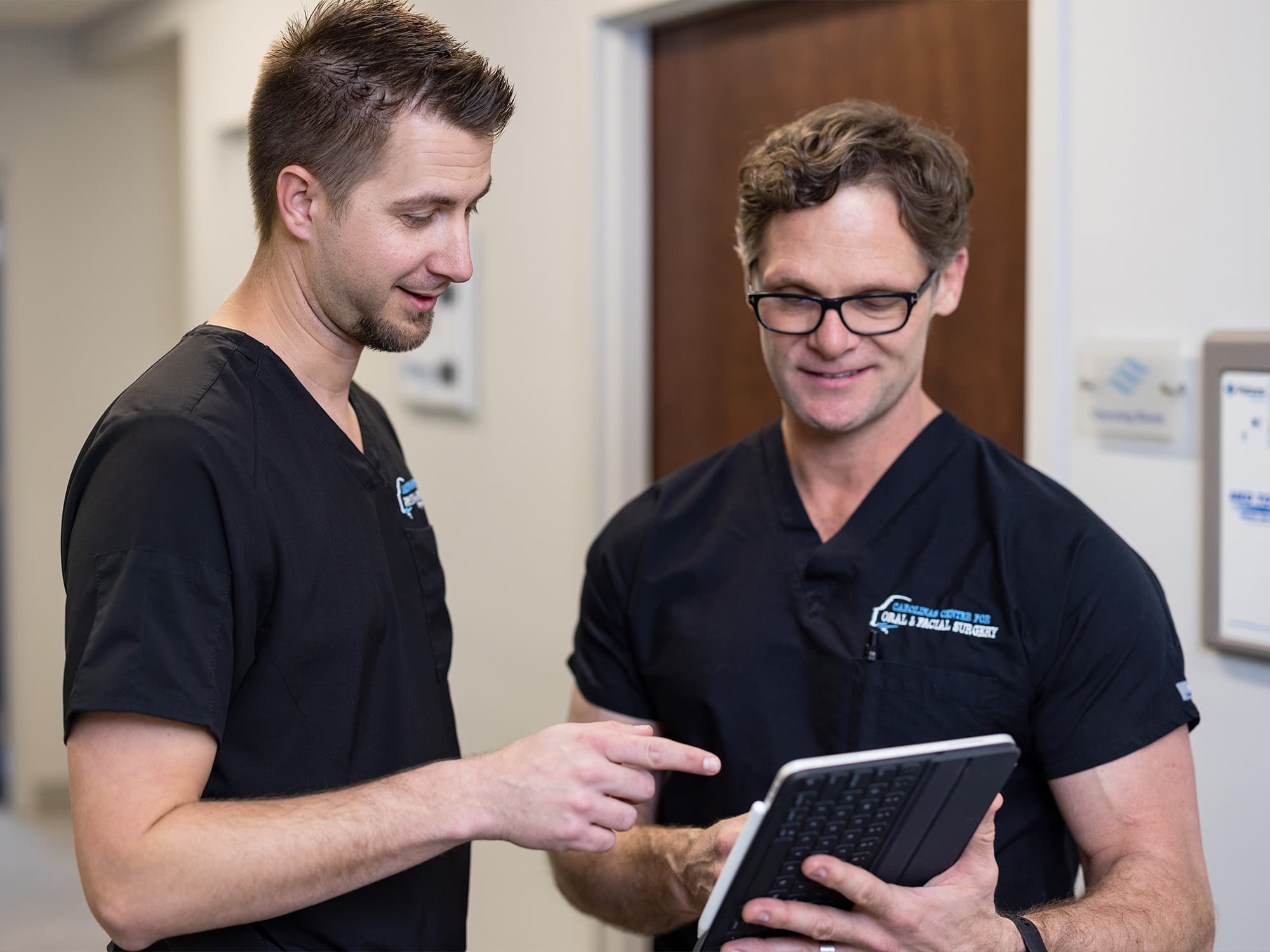 Two people in medical scrubs discuss something on a tablet in a clinical setting, standing near a wall with signs.