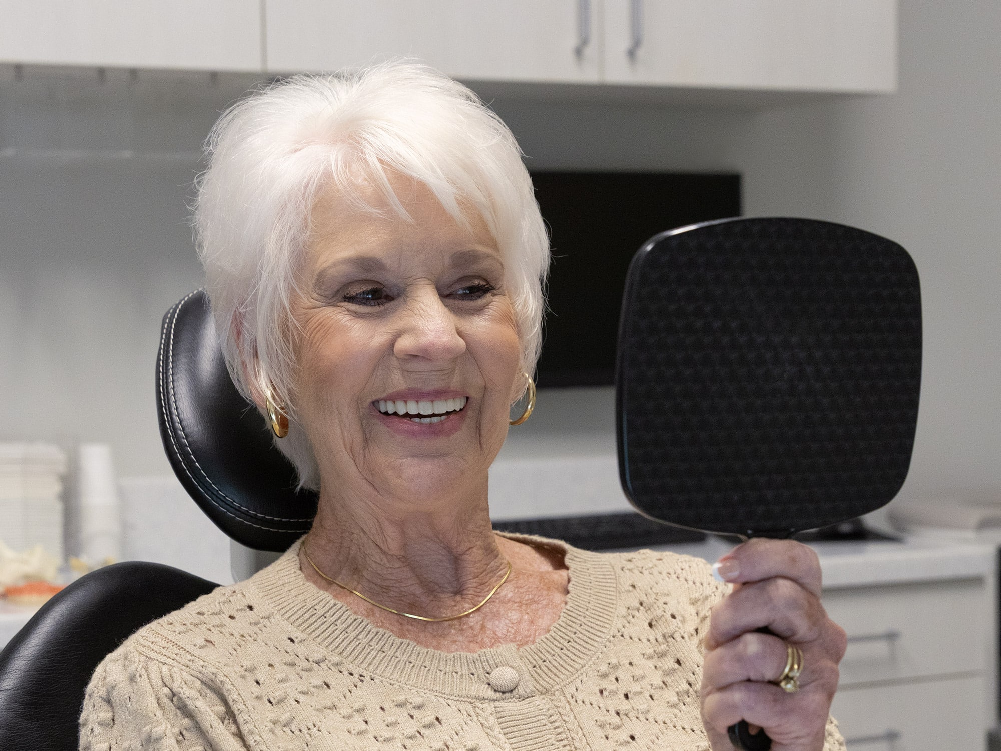 A person with white hair sits happily in a dental chair, holding a mirror, in a modern clinic environment.
