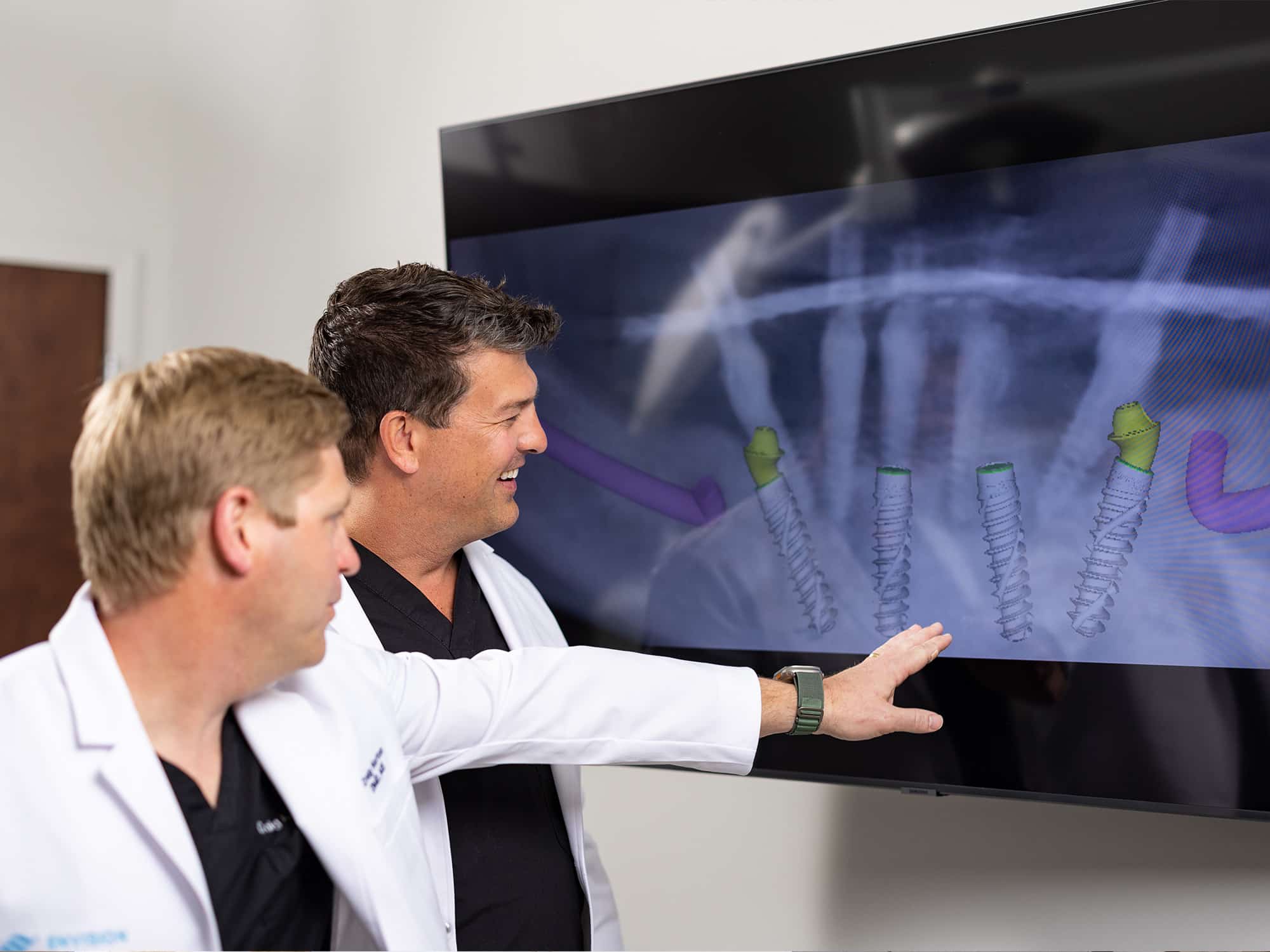 Two people in lab coats examine dental implant images on a screen, discussing and pointing at the diagrams.