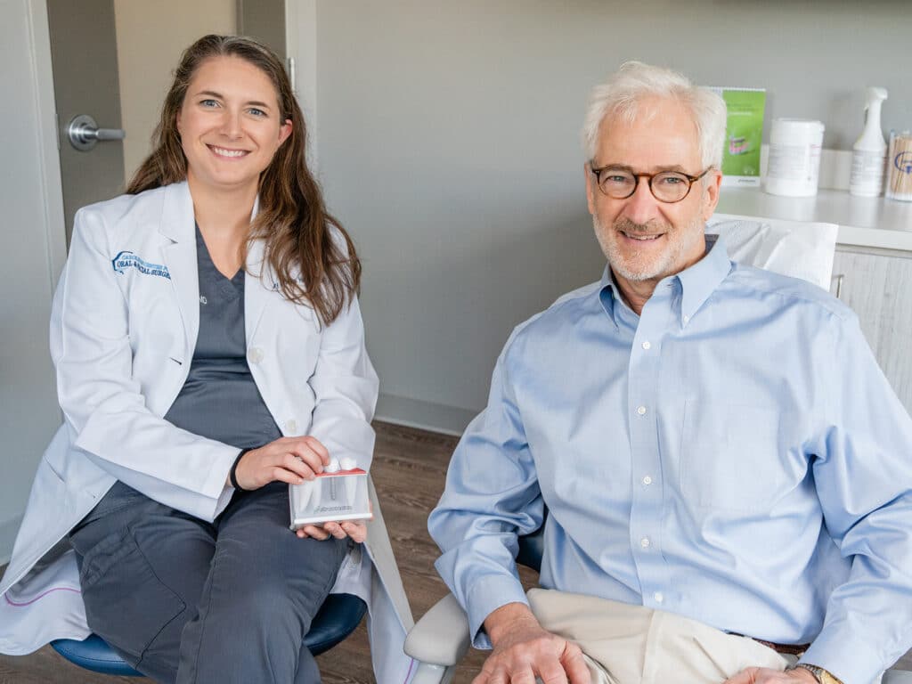Two people in a medical office, one wearing a lab coat, smiling. The setting is professional with medical supplies in the background.