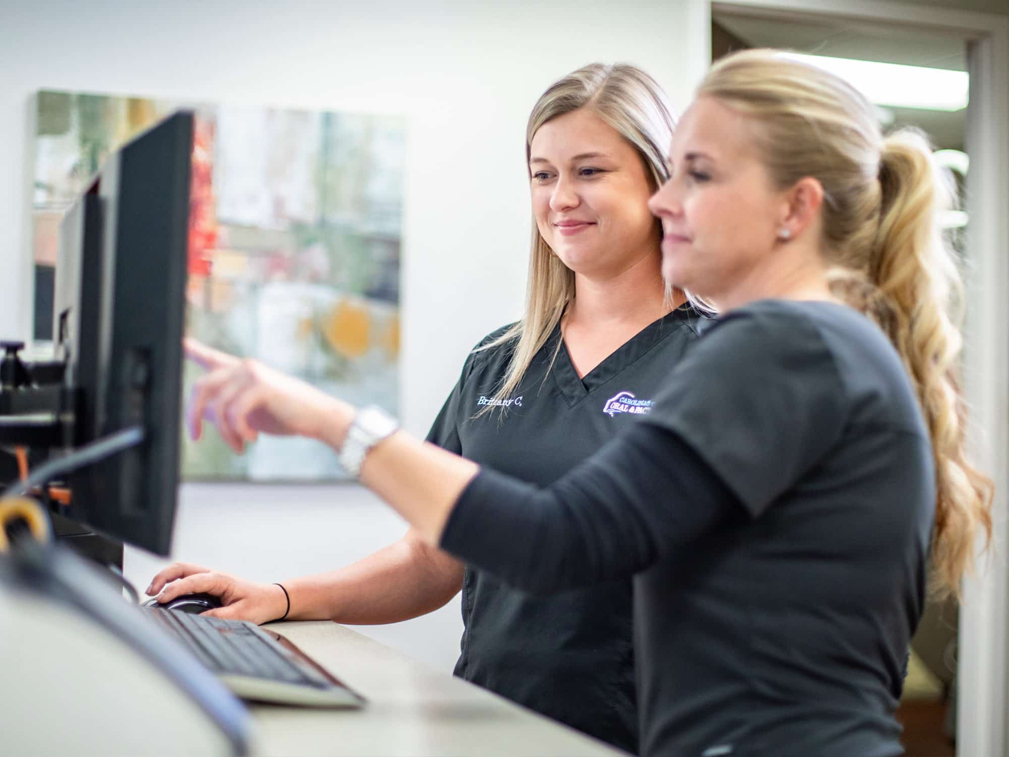 Two people in medical attire work at a computer in an office setting with abstract art on the wall.