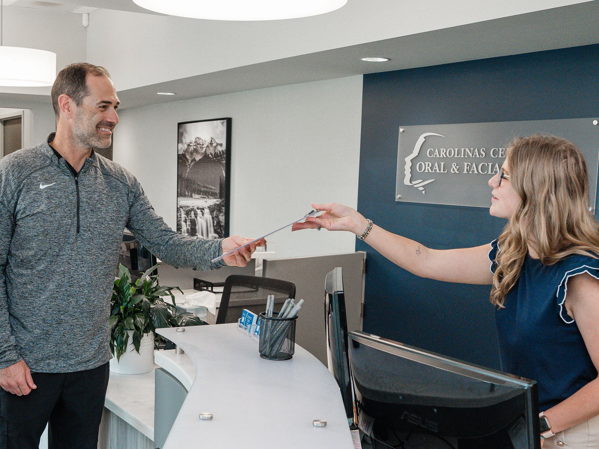 A person at a dental clinic reception hands a document to another person. The backdrop displays a nature photograph and clinic signage.