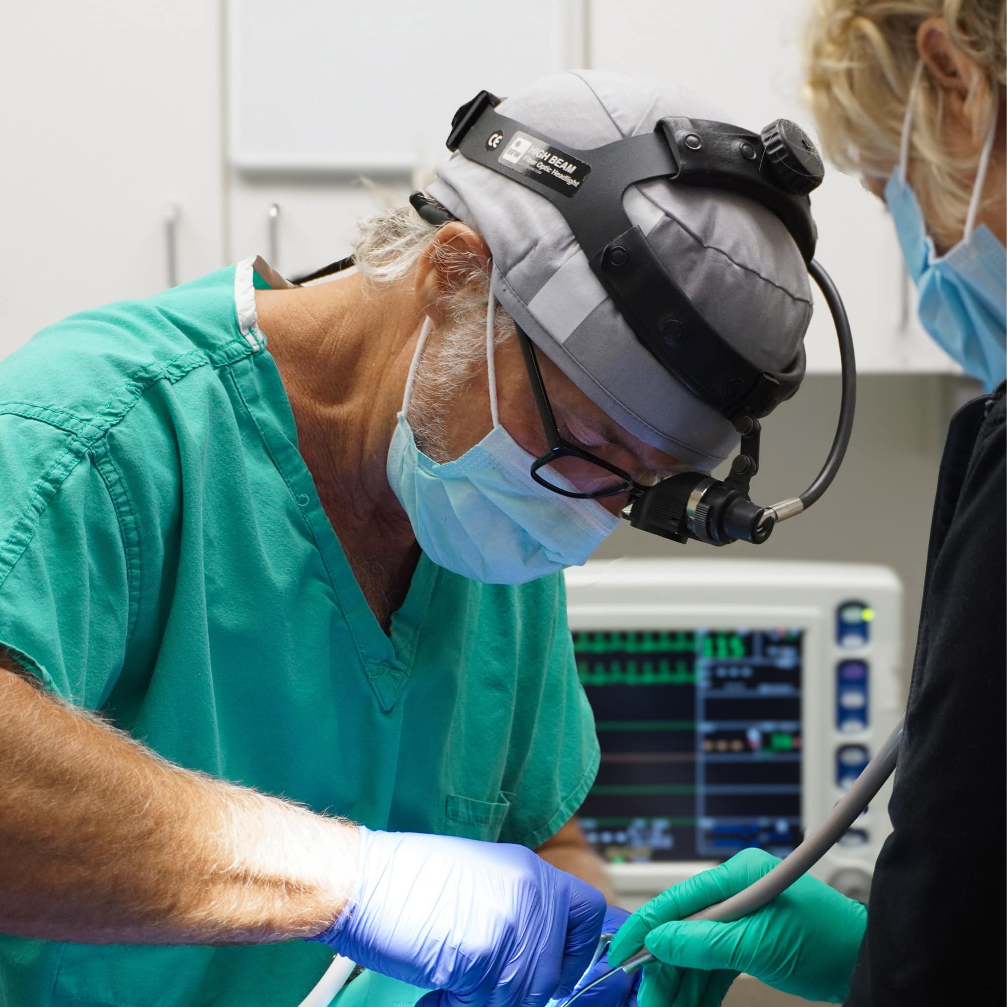 A focused healthcare professional in green scrubs performs surgery, assisted by another. Medical equipment and monitors are visible in the background.