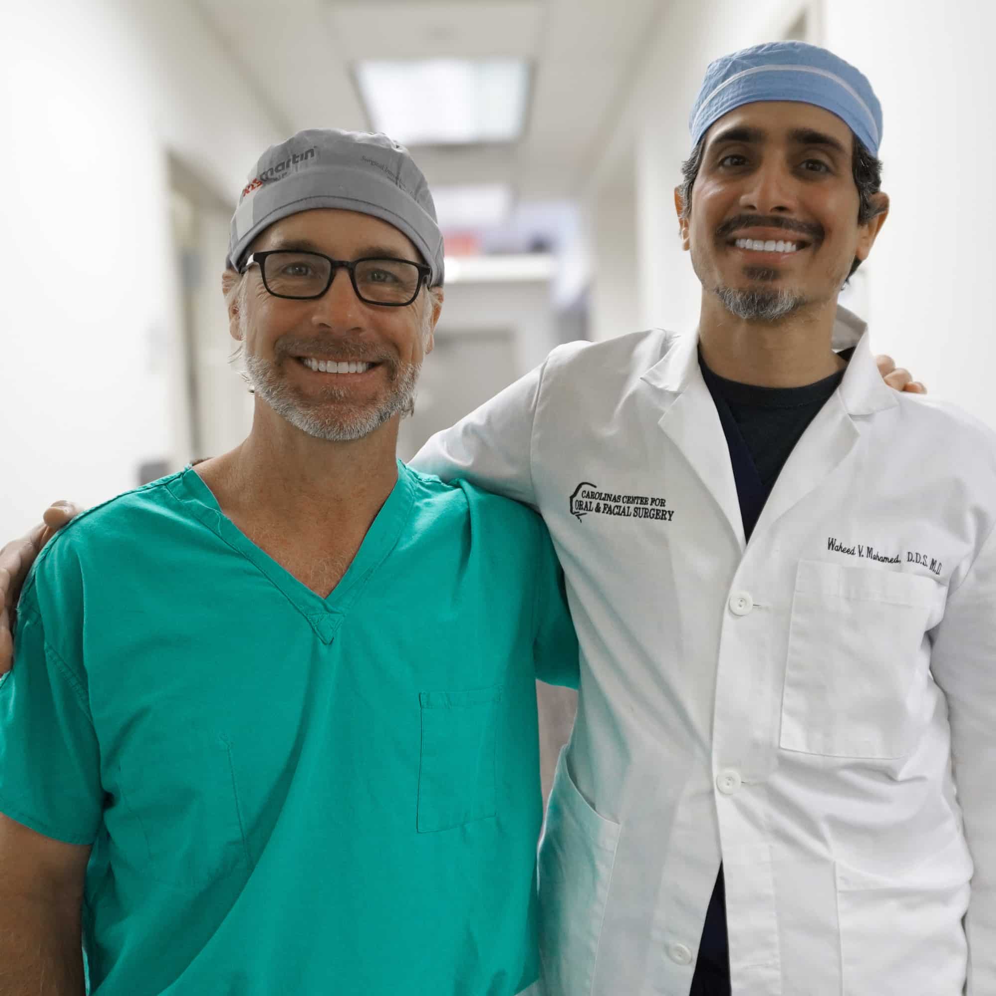 Two people in medical attire stand smiling in a hallway, wearing scrubs and lab coats. The setting appears to be a hospital.