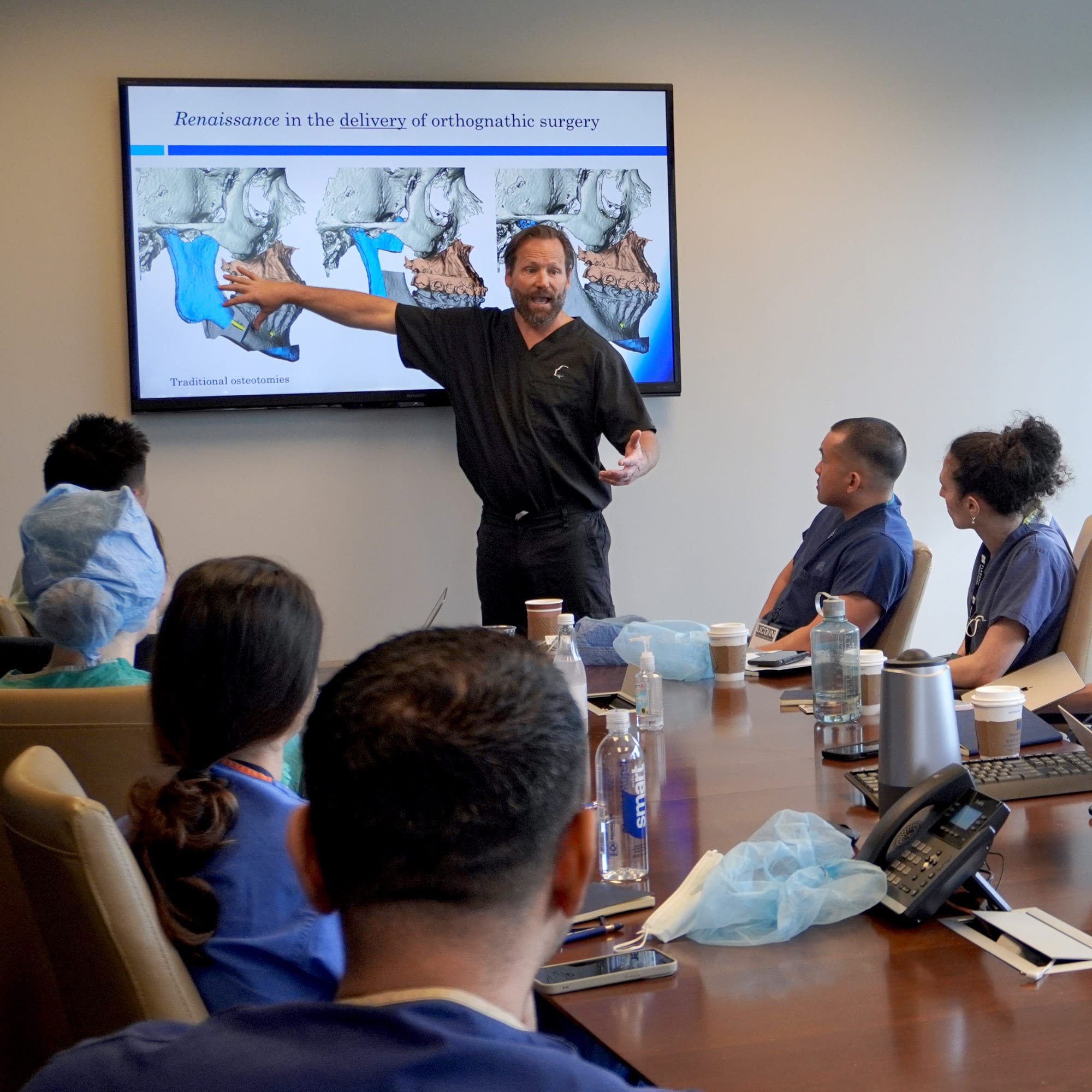 A person presents on orthognathic surgery to five others in a conference room, using a screen displaying anatomical diagrams.