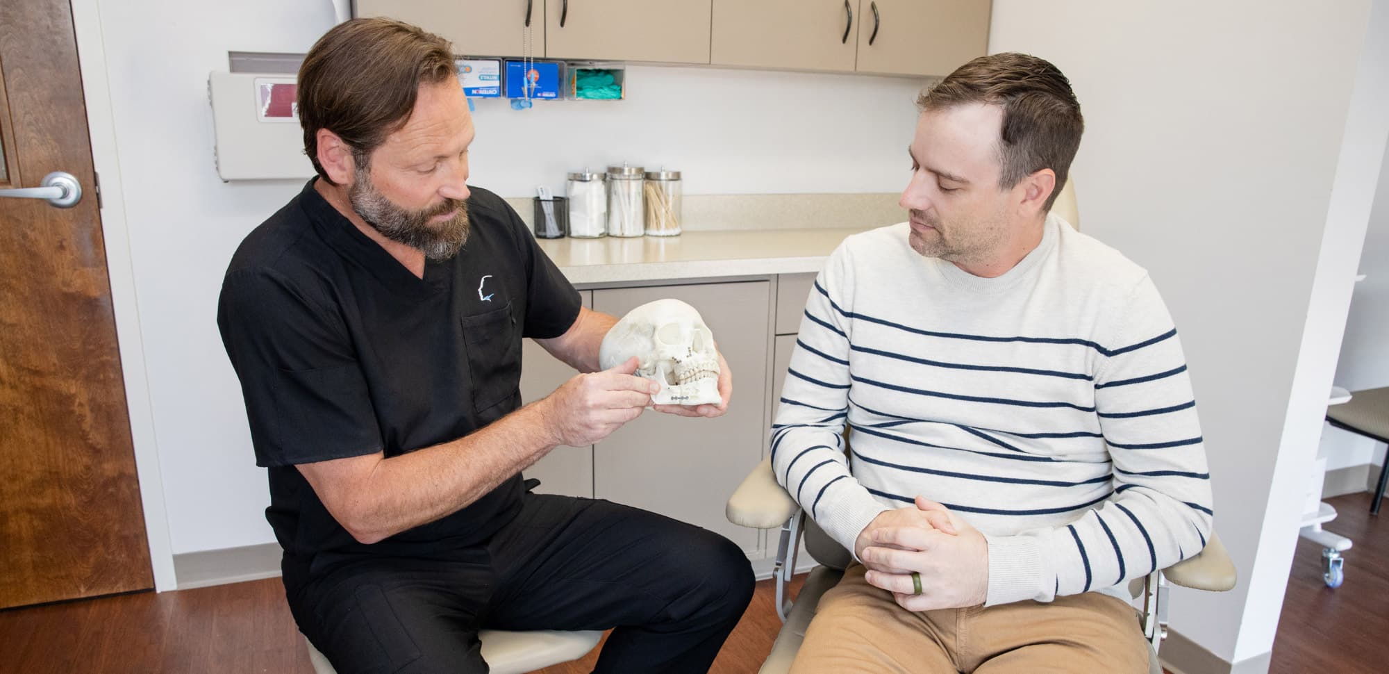 Two people in a medical office. One holds a skull model, explaining something to the other. Cabinets and medical supplies in the background.