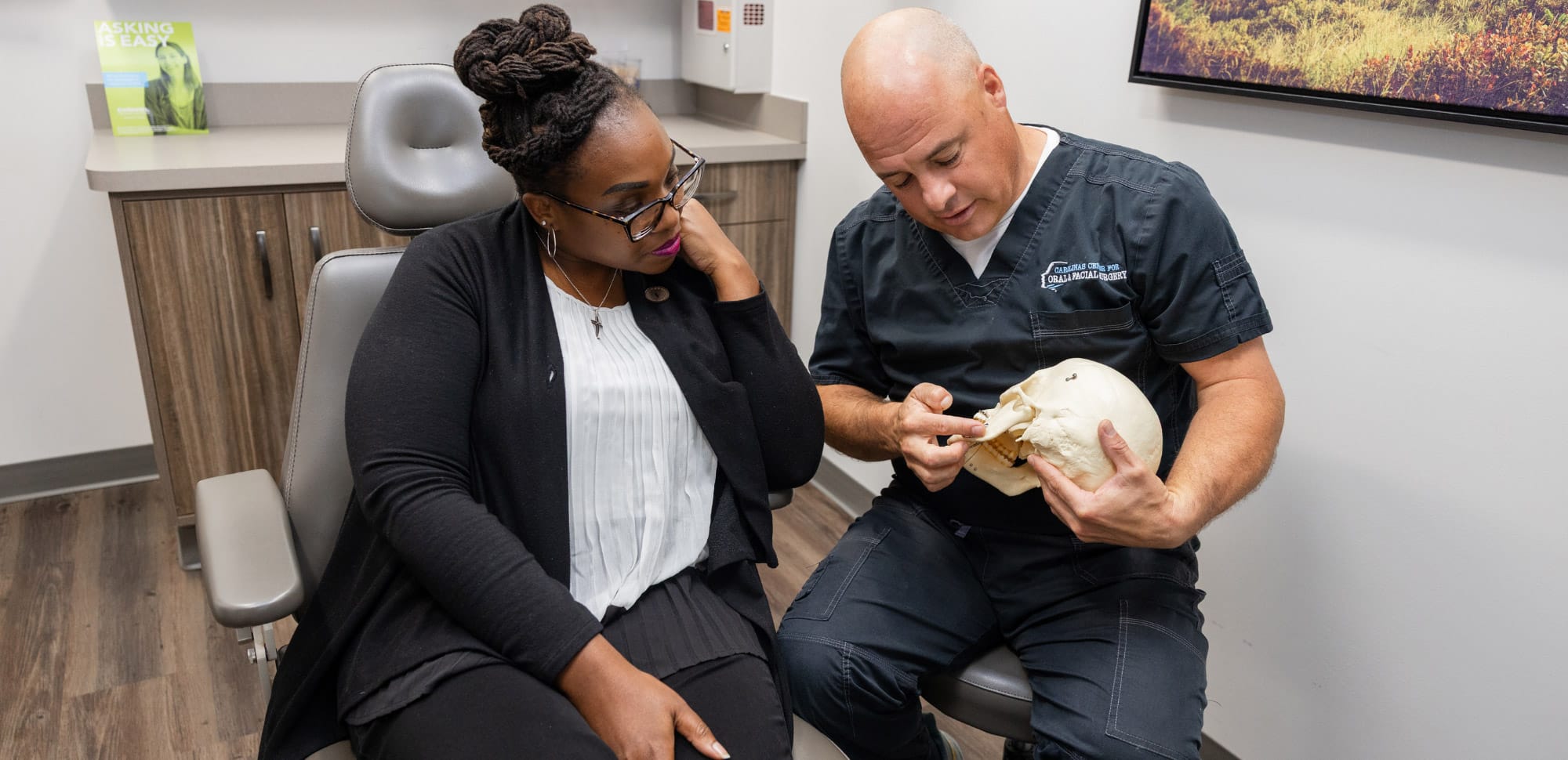 A person in black scrubs discusses a skull model with another seated person in an office setting, emphasizing medical or dental consultation.