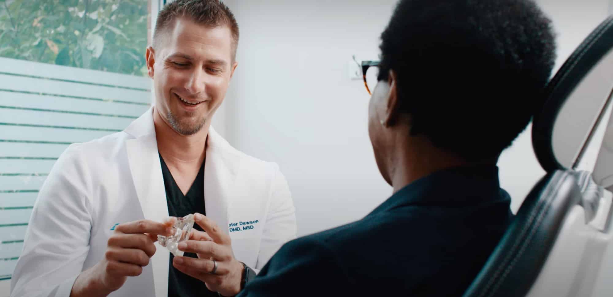 Two people in a consultation; one holding a dental model. Bright setting with a plant outside the window.