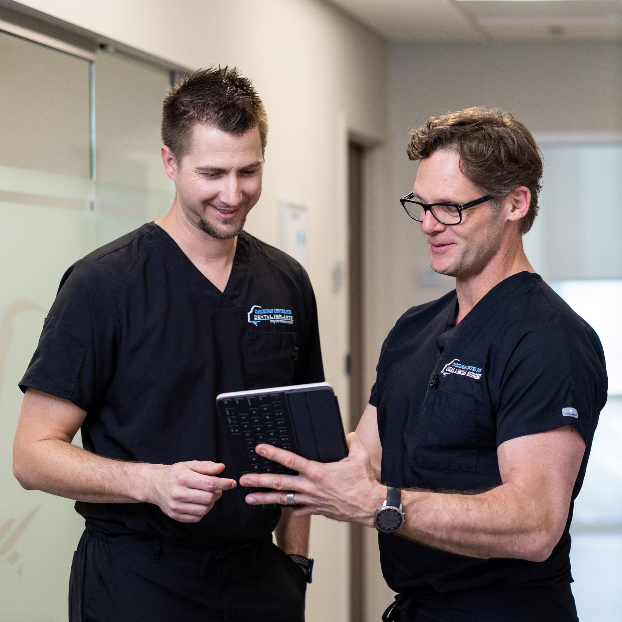 Two people in black scrubs stand in a hallway, discussing and smiling while looking at a tablet device.